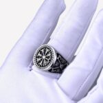 Photo d'une main gantée portant une bague viking argentée avec un symbole de trois triangles et une boussole sur un fond gris foncé