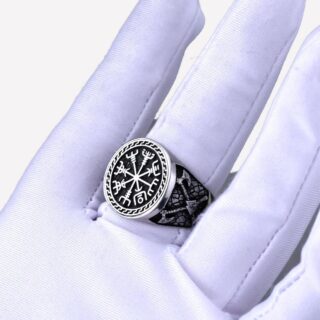 Photo d'une main gantée portant une bague viking argentée avec un symbole de trois triangles et une boussole sur un fond gris foncé