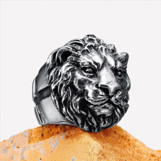 Photo d'une bague tête de lion avec crinière et croix gravée sur le côté posée sur un socle marron clair et fond gris